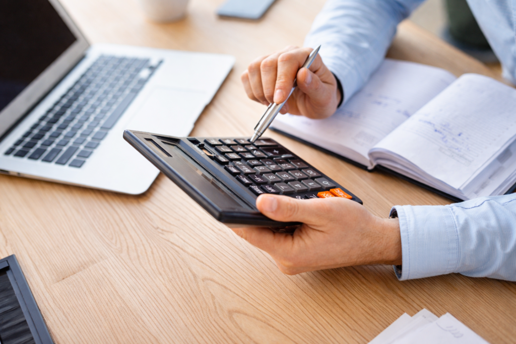 Profissional calculando custos com calculadora sobre mesa de trabalho com notebook e caderno abertos ao lado.